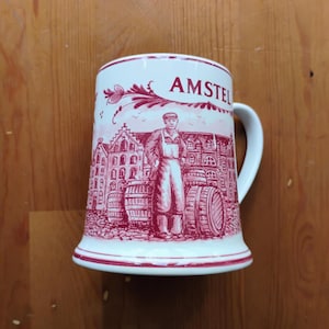 Può includere: Un boccale da birra in ceramica bianca con un'illustrazione stampata in rosso che raffigura una persona in piedi davanti a botti di legno ed edifici tradizionali. Il testo AMSTEL è stampato in rosso in alto. Presenta un manico curvo.