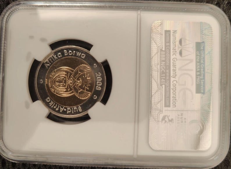 Op de afbeelding: Een Zuid-Afrikaanse munt uit 2009, ingesloten in een rechthoekige NGC beschermende plastic houder. De munt bevat de tekst Afrika Borwa en Suid-Afrika, met een centraal embleem dat een schild en een secretarisvogel weergeeft.