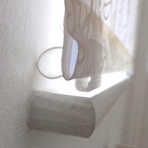 May include: A partially rolled-up window blind with a floral pattern in beige and white. The blind is mounted on a white roller and has a white pull cord. The wall is painted white.