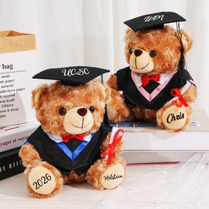 Può includere: Due orsacchiotti marroni con cappelli e vesti di laurea. Un orso ha "UCSC" sul cappello e "2026" e "Holstein" sulle zampe. L'altro orso ha "WPI" sul cappello e "Chris" sulla zampa.