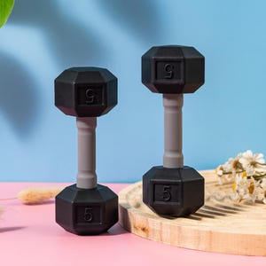 May include: Two black 5-pound dumbbells with gray handles. The dumbbells have a hexagonal shape and the number "5" is printed on each end. The background is a mix of pink and blue.