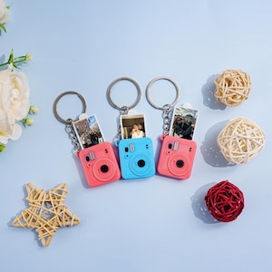 May include: Three miniature camera keychains in coral, blue, and pink, each featuring a metal ring and a small rectangular photograph inserted into the top. The camera designs include a circular lens, a flash, and a shutter button on the front.