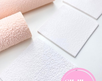 May include: An overhead shot shows several rolls of textured, light pink material and white square tiles. The material appears to be fabric or paper. The white tiles have a similar texture to the pink rolls. The image includes a pink circle with the text "Sibillas cutters".