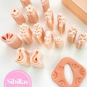 May include: A collection of light peach-colored floral and leaf-shaped cookie cutters. The cutters are arranged with a variety of designs, including flowers, hearts, and leaves. The image also includes a square-shaped cutter with a central oval cutout and floral impressions. The text "Sibillas cutters" is visible.