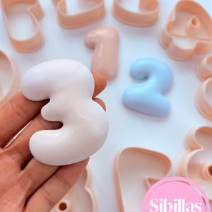 May include: A collection of peach-coloured number-shaped cookie cutters and a single light blue number two cutter. One smooth, white number three shape is held in the foreground. A pink circular logo with the text Sibillas cutters is visible.