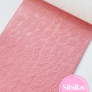 Könnte beinhalten: Eine rosa, strukturierte Teigrolle mit einem sich wiederholenden Blättermuster. Die Teigrolle ist teilweise ausgerollt und zeigt eine weiße, mit Blättern gemusterte Oberfläche. Das Logo "Sibillas cutters" ist in einem rosa Kreis zu sehen.