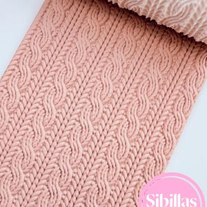May include: A light peach-colored textured sheet featuring a repeating vertical cable knit pattern. At the top, a cylindrical rolling pin with a matching embossed cable knit design is visible. The bottom right corner displays a pink circular logo with the text Sibillas cutters.