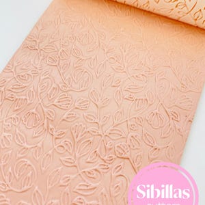 Könnte beinhalten: Eine pfirsichfarbene Teigrolle mit floralem Muster. Das Design zeigt geprägte Blumen und Blätter, wodurch eine strukturierte Oberfläche entsteht. Die Teigrolle ist teilweise ausgerollt und zeigt das detaillierte Muster. Der Markenname "Sibillas cutters" ist sichtbar.