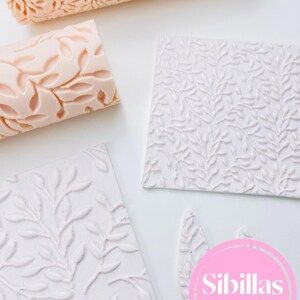 May include: A collection of leaf-patterned baking tools. Includes two rolling pins in a light peach color and several white textured square and leaf-shaped cutters. The tools are arranged on a white surface. The brand name "Sibillas cutters" is visible.