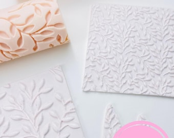 May include: A collection of leaf-patterned baking tools. Includes two rolling pins in a light peach color and several white textured square and leaf-shaped cutters. The tools are arranged on a white surface. The brand name "Sibillas cutters" is visible.