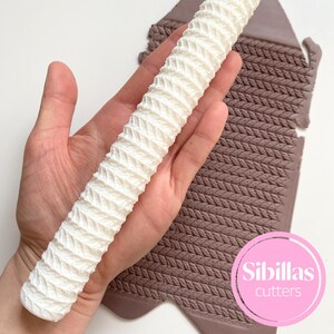 May include: A white, textured rolling pin with a knitted pattern is held in a hand. A brown, textured mat with a matching knitted pattern is in the background. The rolling pin and mat are likely for baking or crafting.