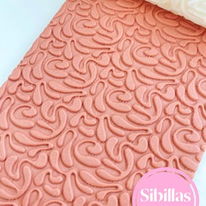 Könnte beinhalten: Eine rosa Teigrolle mit einem strukturierten Muster aus wirbelnden Formen. Die Teigrolle ist teilweise auf einer passenden rosa Oberfläche ausgerollt, wodurch das geprägte Design sichtbar wird. Der Markenname "Sibillas cutters" ist in einem rosa Kreis zu sehen.
