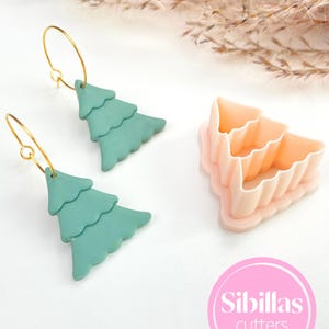 Peut inclure: Boucles d'oreilles sapin de Noël turquoise avec des cerceaux dorés et un emporte-pièce rose et orange. L'image comprend le texte "Sibillas cutters" dans un cercle rose.