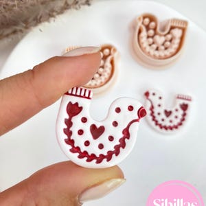 May include: A white and red cookie cutter in the shape of a bird is held between fingers. The bird design features a heart and floral pattern. Two additional cookie cutters are visible in the background.