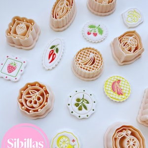 Könnte beinhalten: Eine Sammlung beigefarbener Ausstecher in verschiedenen Formen, darunter Früchte und Blumen. Mehrere dekorierte Kekse mit Fruchtmotiven sind ebenfalls vorhanden. Das Bild enthält den Text "Sibillas cutters" in einem rosa Kreis.