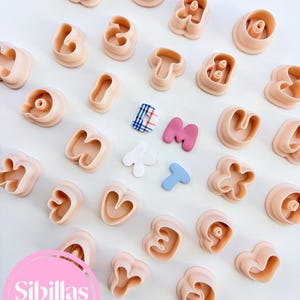 May include: A collection of peach-colored plastic alphabet cookie cutters arranged on a white surface. Three small letter shapes in plaid, pink, and blue are placed in the center. A pink circular logo with the text Sibillas cutters is visible.