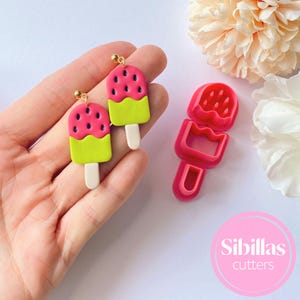 Pode incluir: Brincos em forma de melancia com pinos dourados e um conjunto de cortadores de argila polimérica a condizer. Os brincos apresentam um topo rosa com detalhes de sementes pretas, um meio verde e um pau branco. O conjunto de cortadores é rosa.