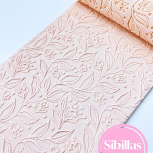 Może przedstawiać: Jasnoróżowy wałek do ciasta z kwiatowym wzorem, częściowo rozwinięty. Wzór zawiera liście, kwiaty i kropki. Wałek wykonany jest z solidnego materiału i nadaje się do pieczenia i rękodzieła. Widoczna nazwa marki.