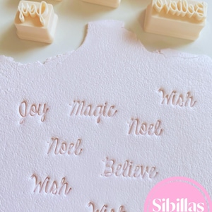 Peut inclure: Un ensemble de cinq tampons rectangulaires blanc cassé avec des lettres cursives en relief, incluant les mots Joy, Magic, Noel, Believe et Wish. Sous les tampons, une surface blanche texturée présente les mêmes mots embossés.