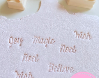 May include: A set of five off-white rectangular stamps with raised cursive lettering, including the words Joy, Magic, Noel, Believe, and Wish. Below the stamps, a white textured surface features the same words embossed into the material.