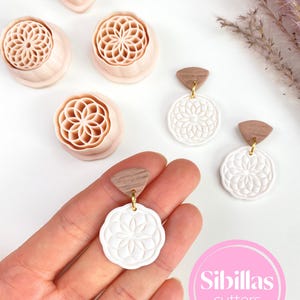 Op de afbeelding: Een verzameling lichtroze polymeerklei-uitsteekvormen en witte oorbellen met een bloemmotief. De oorbellen hebben houten bovenkanten en goudkleurige haken. De afbeelding bevat de tekst "Sibillas cutters" in een roze cirkel.