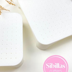 May include: Two white, rectangular cutters with rounded corners and small, evenly spaced holes. The cutters have a slightly raised edge and are set against a white background. The image includes a pink circle with the text "Sibillas cutters".