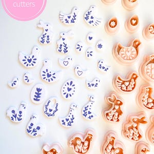 May include: A collection of pink cookie cutters in various shapes, including birds, rabbits, and eggs, alongside white cookies decorated with blue floral patterns. The image includes the text "Sibillas cutters" in a pink circle.