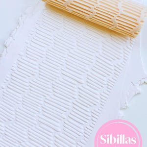 May include: A beige textured rolling pin with a repeating oval pattern, used to imprint a design on white clay. The clay is embossed with the same pattern. The image includes a pink circle with the text "Sibillas cutters".
