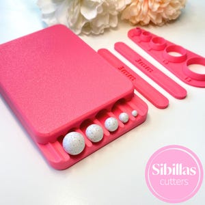 Puede incluir: Un juego de herramientas de artesanía rosa brillante con varias formas y tamaños. El juego incluye una base rectangular con ranuras curvas que sostienen esferas blancas moteadas. Piezas adicionales incluyen reglas marcadas "3mm" y cortadores circulares. El logotipo "Sibillas cutters" es visible.