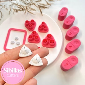 May include: A collection of pink and white craft cutters and tools. The image features several pink, triangular cutters with three circular holes, along with numbered pink rectangular pieces. Two white, triangular cutters are held in a hand, and the text "Sibillas cutters" is visible.