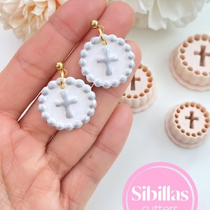 May include: White and gray cross earrings with gold-colored hooks. The earrings have a circular design with a cross in the center and a beaded border. The image also shows cookie cutters with a cross design and the text "Sibillas cutters".