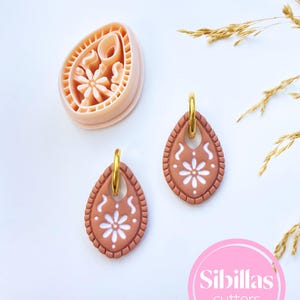 May include: A set of terracotta-colored teardrop earrings with a white floral design and gold hoops. A matching cutter is also visible. The earrings have a textured border and are displayed on a white surface. The image also includes the text "Sibillas cutters".