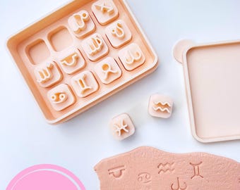 May include: A set of light peach-colored zodiac sign cookie cutters with a matching tray and lid. The cutters are arranged in a tray, with a few loose. A piece of clay displays stamped zodiac symbols. The logo "Sibillas cutters" is in a pink circle.