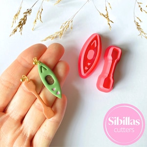 Op de afbeelding: Een paar polymer clay oorbellen met een groene kano met roze hartjes en een bruin peddel. De oorbellen worden getoond naast hun rode siliconen mallen, die worden gebruikt om de oorbellen te maken.