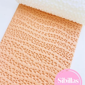 May include: A white textured rolling pin partially rolled over a light orange sheet with a repeating pattern of small ovals. The sheet has a textured surface. The image includes a pink circle with the text "Sibillas cutters".