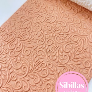 Könnte beinhalten: Ein terrakottafarbener, strukturierter Nudelholz und eine Fondantplatte mit einem geprägten floralen Design. Das Nudelholz ist weiß mit einem passenden Blumenmuster. Der rosa Kreis mit dem Text "Sibillas cutters" befindet sich unten rechts.