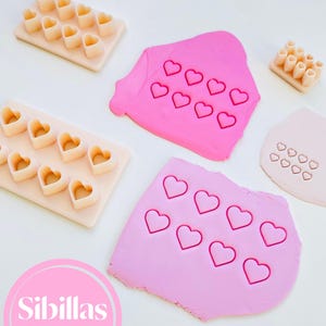 Può includere: Vista dall'alto di taglia biscotti a forma di cuore e argilla rosa. I taglia biscotti sono disposti in vassoi, mentre l'argilla ha impronte di cuori. L'immagine include il testo "Sibillas cutters" in un cerchio rosa.