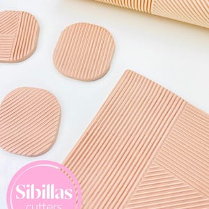 Könnte beinhalten: Eine Sammlung von strukturierten, hellpfirsichfarbenen Ton-Ausstechern und einer Teigrolle. Die Ausstecher haben verschiedene Formen, darunter Kreise und geometrische Designs. Die Teigrolle hat ein geripptes Muster. Der Markenname "Sibillas cutters" ist sichtbar.