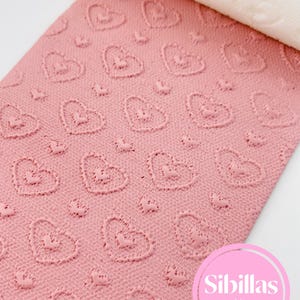 May include: A pink rolling pin with a heart-shaped pattern. The rolling pin is partially unrolled, showing a textured surface with repeating heart shapes. The brand name "Sibillas cutters" is visible in a pink circle.