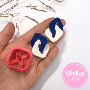 Könnte beinhalten: Zwei Paar quadratische Ohrringe in Blau und Weiß mit Sprenkelmuster und verbundenem Design. Ein rosa Ausstecher mit ovaler Form ist ebenfalls abgebildet. Die Ohrringe sind handgefertigt und haben einen modernen, geometrischen Stil.