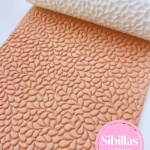 May include: A close-up of a textured, light brown fondant sheet with a leaf pattern, alongside a white rolling pin with a matching design. The image includes a pink and white logo that says "Sibillas cutters".