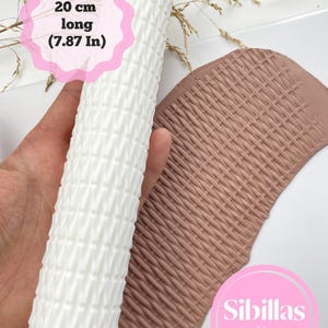 Pode incluir: Um rolo branco texturizado, com 20 cm de comprimento, com um padrão tecido, ao lado de um pedaço de argila marrom com uma textura tecida correspondente. A imagem inclui o texto "Sibillas cutters".