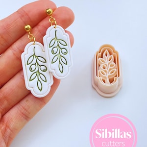 Peut inclure: Boucles d'oreilles blanches et dorées avec un motif de branche d'olivier vert. Les boucles d'oreilles sont rectangulaires avec des bords arrondis et des clous dorés. Un emporte-pièce assorti en forme de branche d'olivier est également visible.