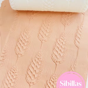 May include: A white cylindrical rolling pin with a wheat stalk pattern embossed on a flat, light peach-coloured clay surface. The rolling pin creates a repeating texture of wheat ears. A pink circular logo with the text Sibillas cutters is visible.