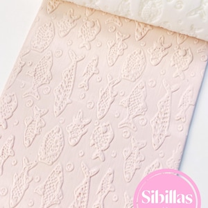 May include: A white cylindrical rolling pin with a raised fish pattern is shown pressing into a light pink surface, creating an embossed design of various fish and small circles. A pink circular logo reads Sibillas cutters.
