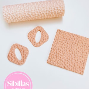 May include: A selection of light peach polymer clay cutters. The set includes a textured rolling pin, two square cutters with oval cutouts, and a textured square. The Sibillas Cutters logo is in the lower left corner.