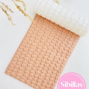 Puede incluir: Un rodillo cilíndrico blanco con un patrón repetitivo en forma de corazón, colocado sobre una lámina rectangular de arcilla de color tostado claro grabada con el mismo diseño. El texto Sibillas cutters es visible en un círculo rosa.