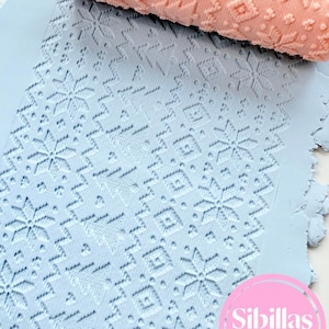 Könnte beinhalten: Ein pfirsichfarbenes, gemustertes Nudelholz liegt auf einer hellblauen Tonplatte. Das Nudelholz weist ein sich wiederholendes winterliches Design aus Schneeflocken und geometrischen Formen auf, das in den Ton geprägt ist. Ein rosa kreisförmiges Logo zeigt den Text Sibillas cutters.