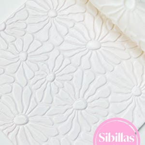 Op de afbeelding: Een witte deegroller met een verhoogd bloemenpatroon en een bijpassende witte plaat materiaal met hetzelfde madeliefjespatroon. Een cirkelvormig roze logo in de rechterbenedenhoek bevat de witte tekst Sibillas cutters.