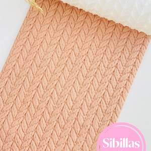 May include: A peach-colored, textured rolling pin with a cable knit pattern. The rolling pin is partially unrolled, revealing a white, textured surface. The image includes the text "Sibillas cutters" in a pink circle.